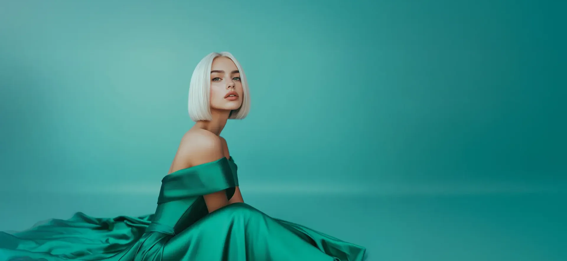 Kobieta w zielonej, satynowej sukni siedząca na tle jednolitego, turkusowego tła. Ma krótkie, proste, jasne włosy i patrzy wprost, wyrażając subtelną elegancję.